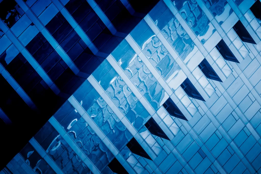 Blue-tinted, diagonal abstract shot of a modern building's glass facade, showing reflections of the sky and other structures.